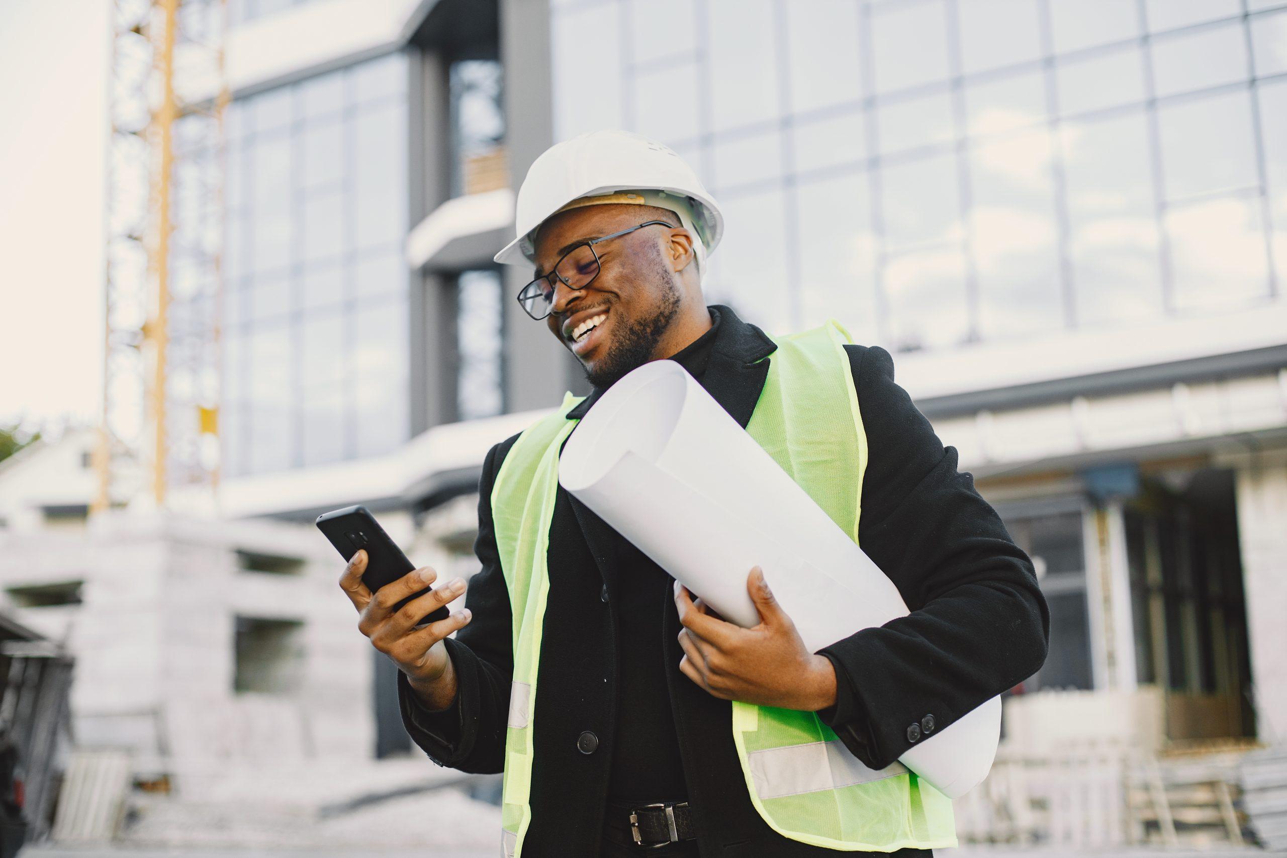 Young black race man with blueprint standing near glass building. Talking by phone.